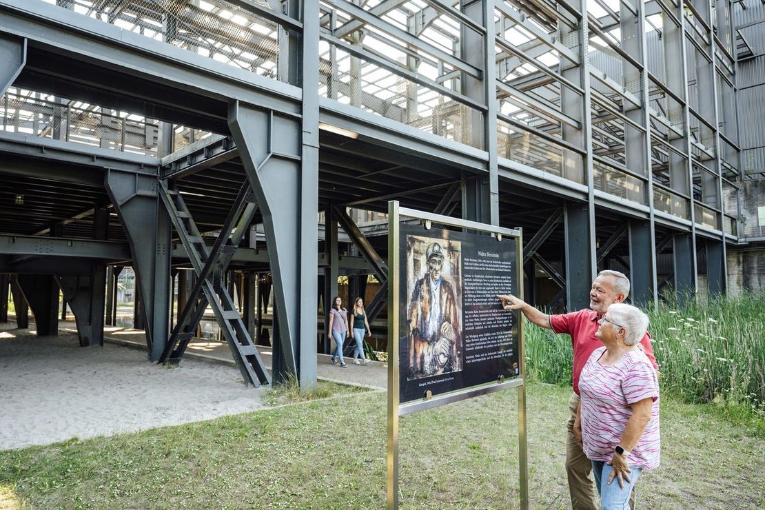 Walter Bernsteinausstellung. Ein Paar steht vor einer Bildtafel der Walter Bernstein Ausstellung im Wassergarten. Im Hintergrund ist die Sieberei des ehemaligen Bergwerks Reden zu sehen